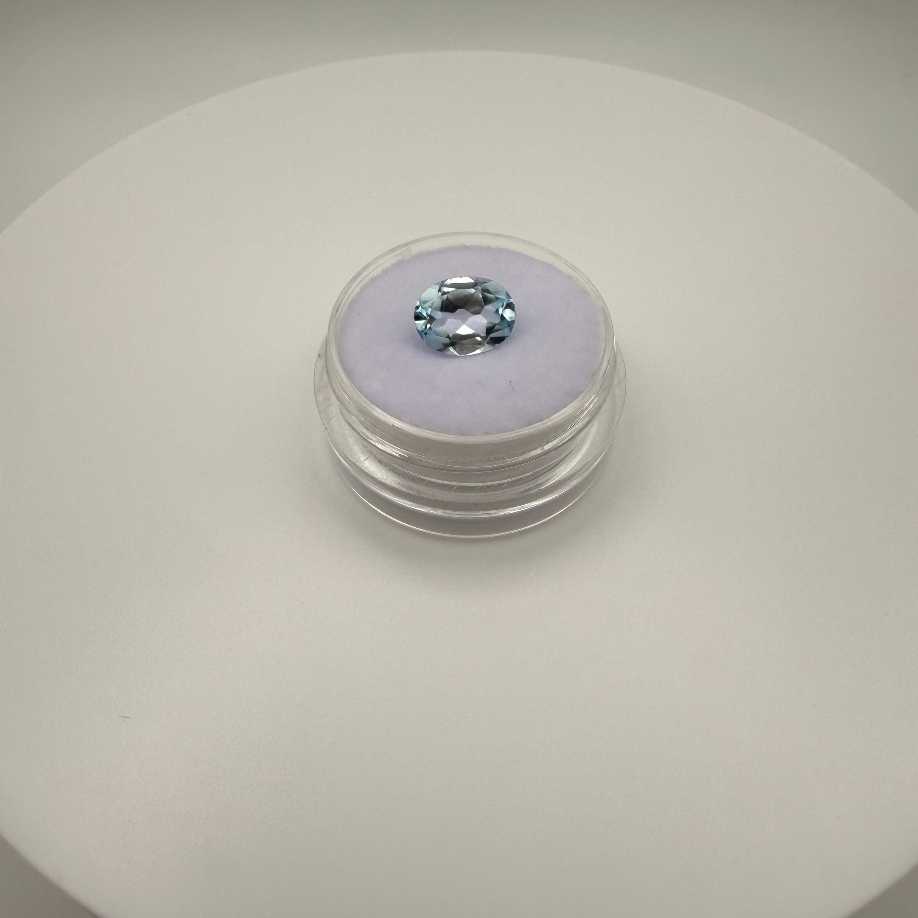 Top view of a 2.95ct oval Swiss Blue Topaz gemstone on a soft lavender cushion in a clear display case.