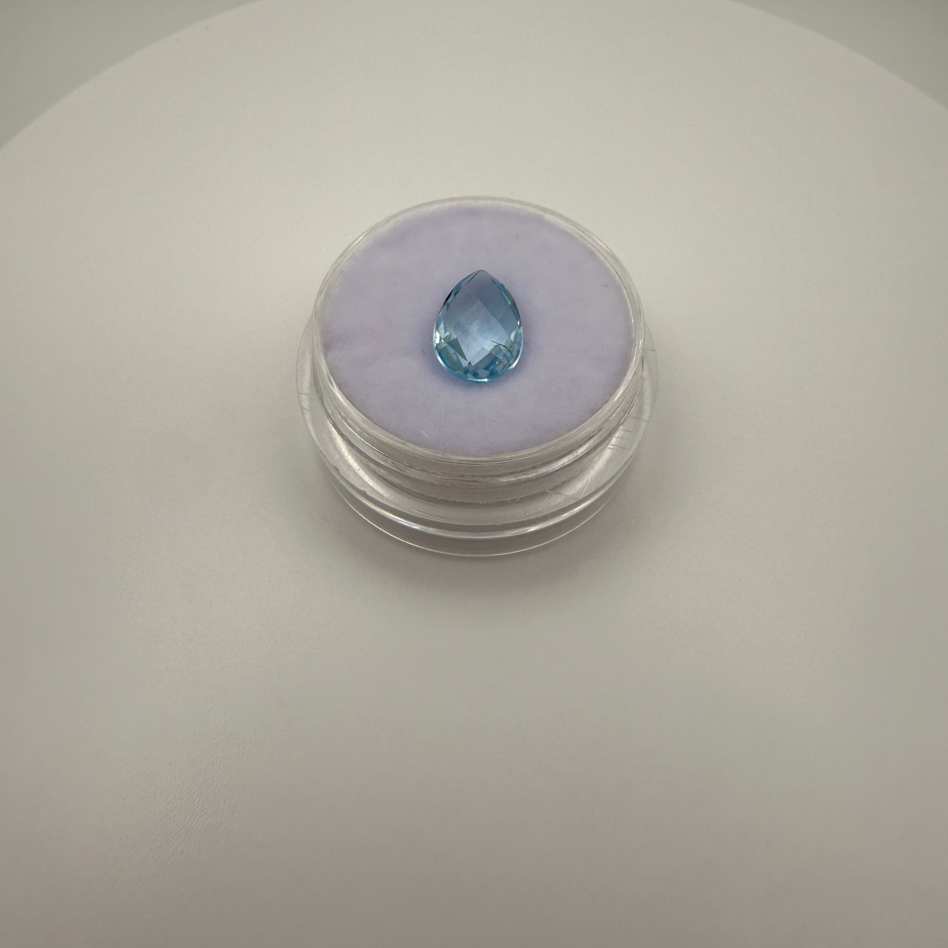 top down view of 2.0ct Swiss Blue Topaz pear faceted gemstone on purple cushion in clear gem holder on white background