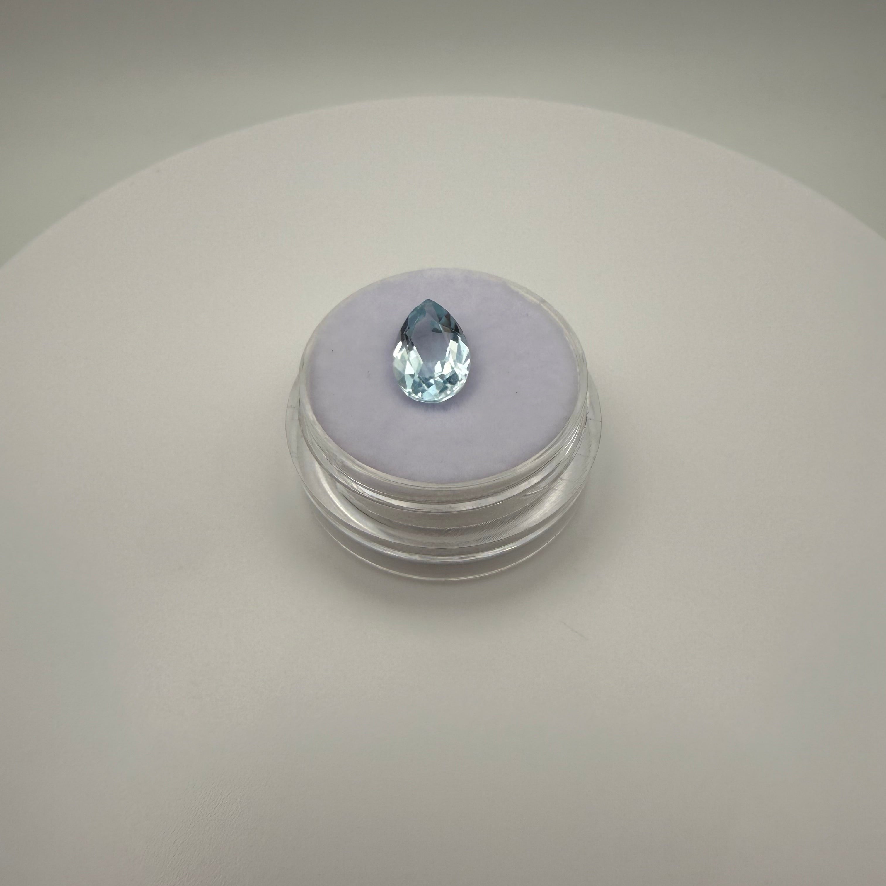 Top-down view of a 2.25ct Swiss Blue Topaz pear faceted gemstone on a soft display surface in a clear gem holder.
