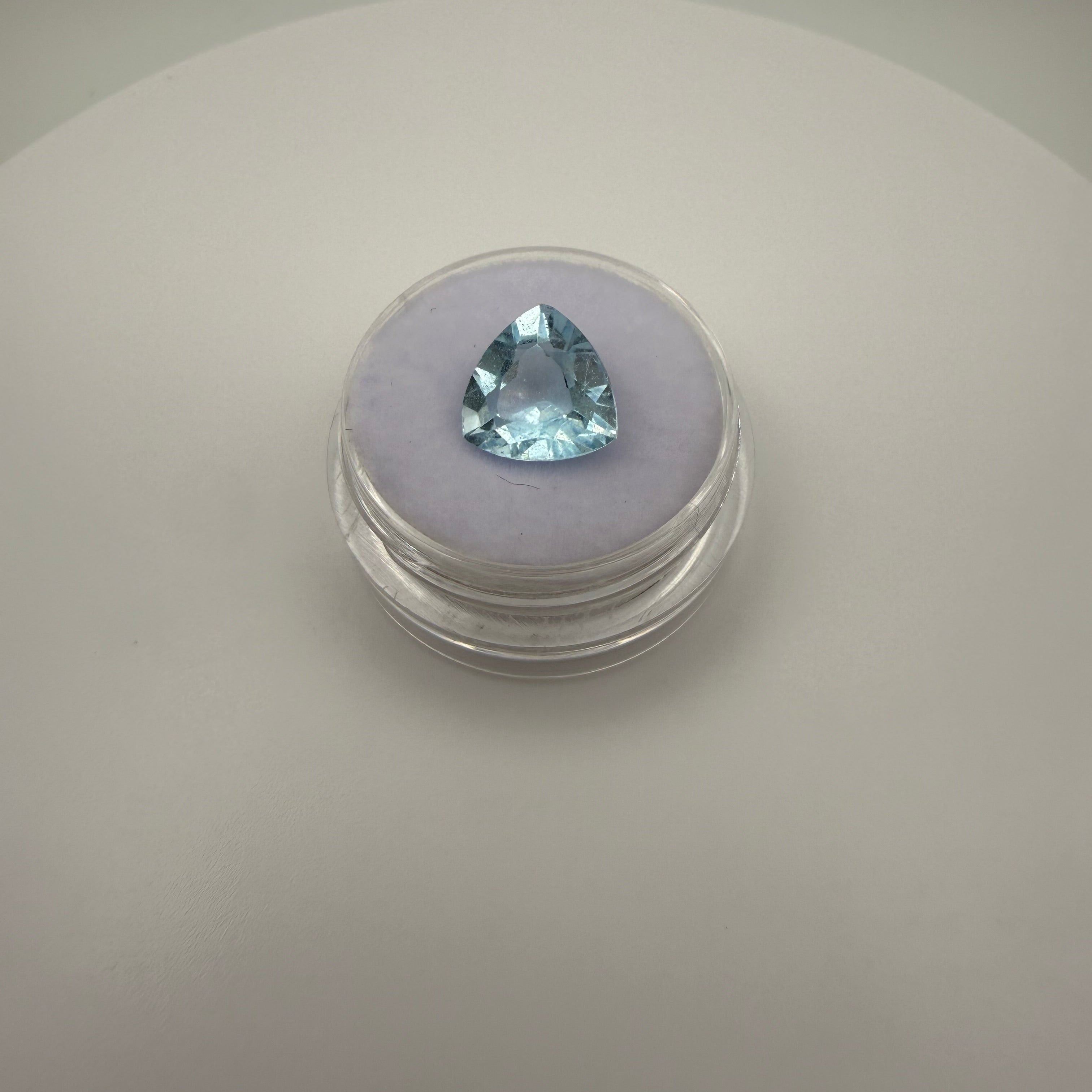 triangular faceted 3.6ct Swiss Blue Topaz gemstone on a purple cushion in a clear gem holder