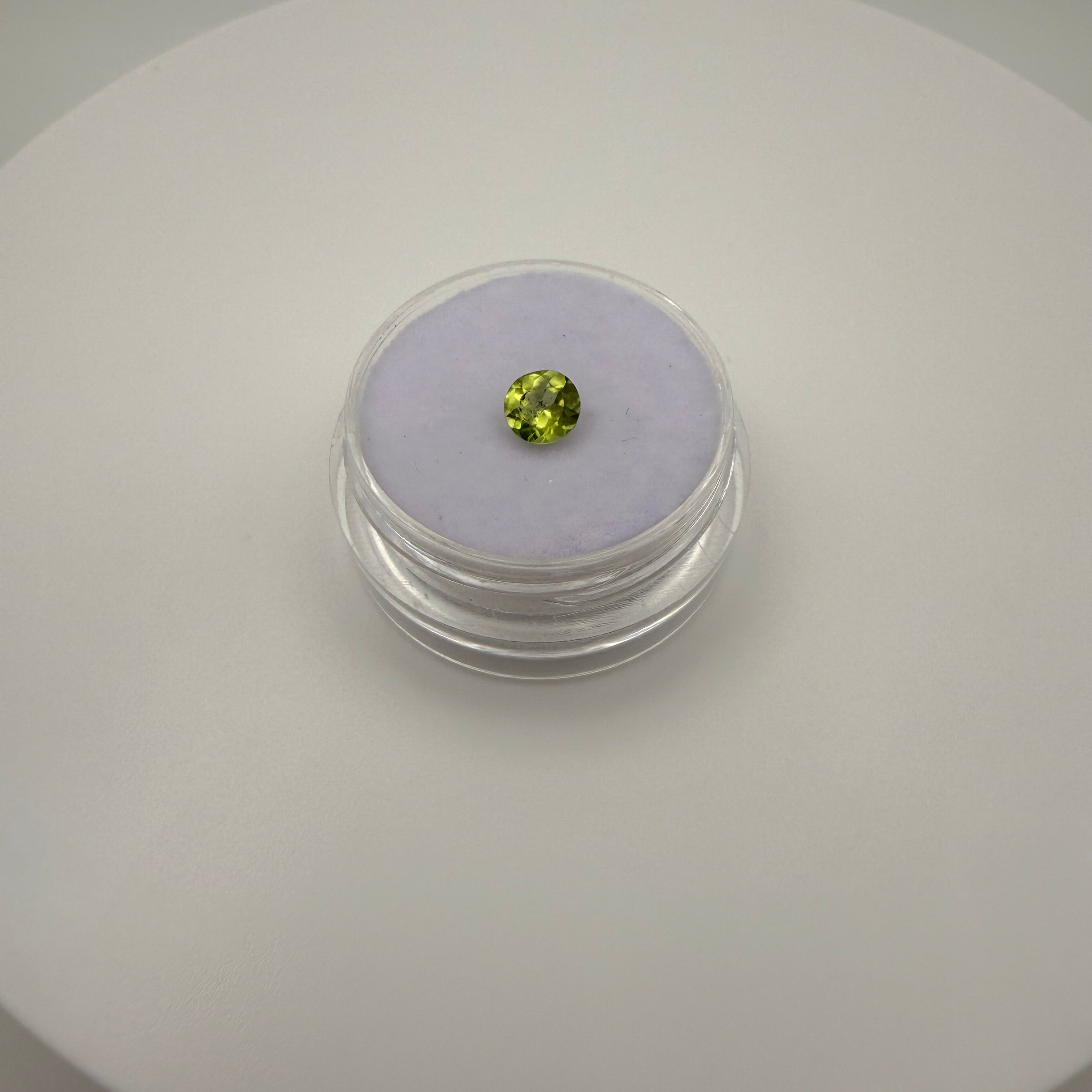 Macro top-down view of a 0.45ct yellow-green peridot round faceted gemstone on a soft purple cushion in a clear gem holder.