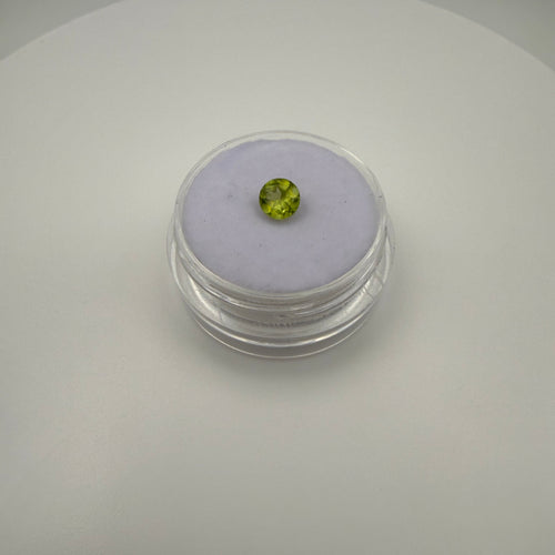 Round faceted 0.45ct yellow-green peridot gemstone displayed in a clear holder on soft surface