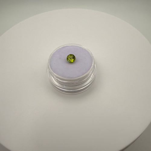 Macro top down view of 0.5ct yellow-green peridot round faceted gemstone on a light purple cushion inside a clear holder.