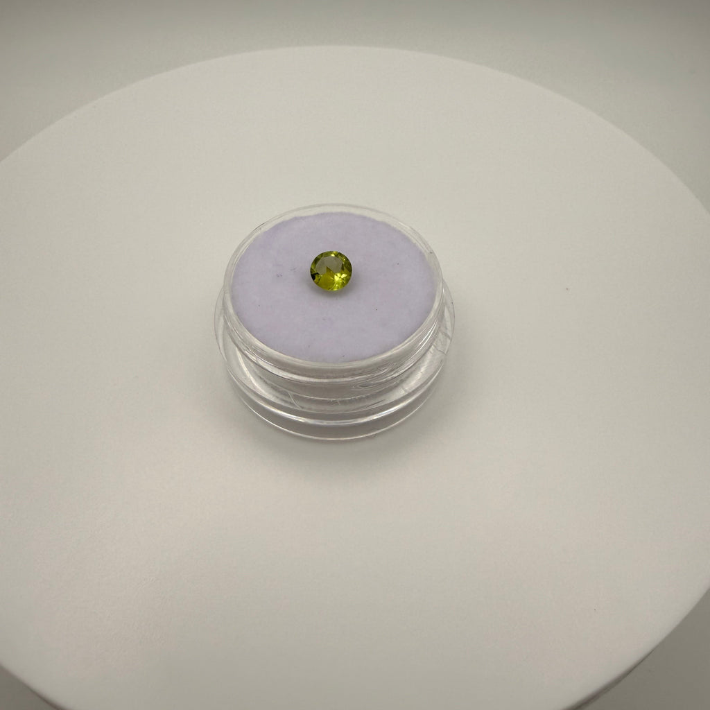 Macro top-down view of a 0.45ct yellow-green peridot round faceted gemstone on a soft purple cushion in a clear display holder.