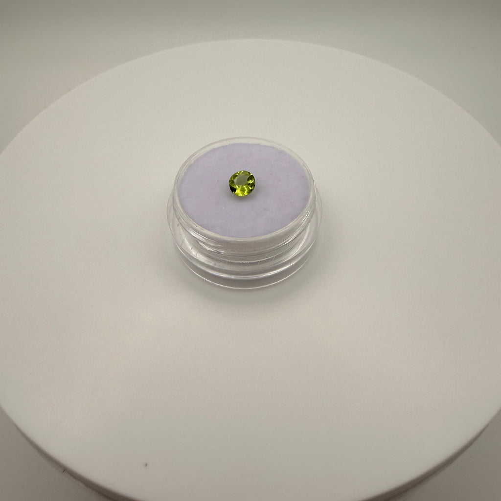 Macro view of a 0.45ct yellow-green peridot round faceted gemstone in a clear gem holder on a white circular surface.