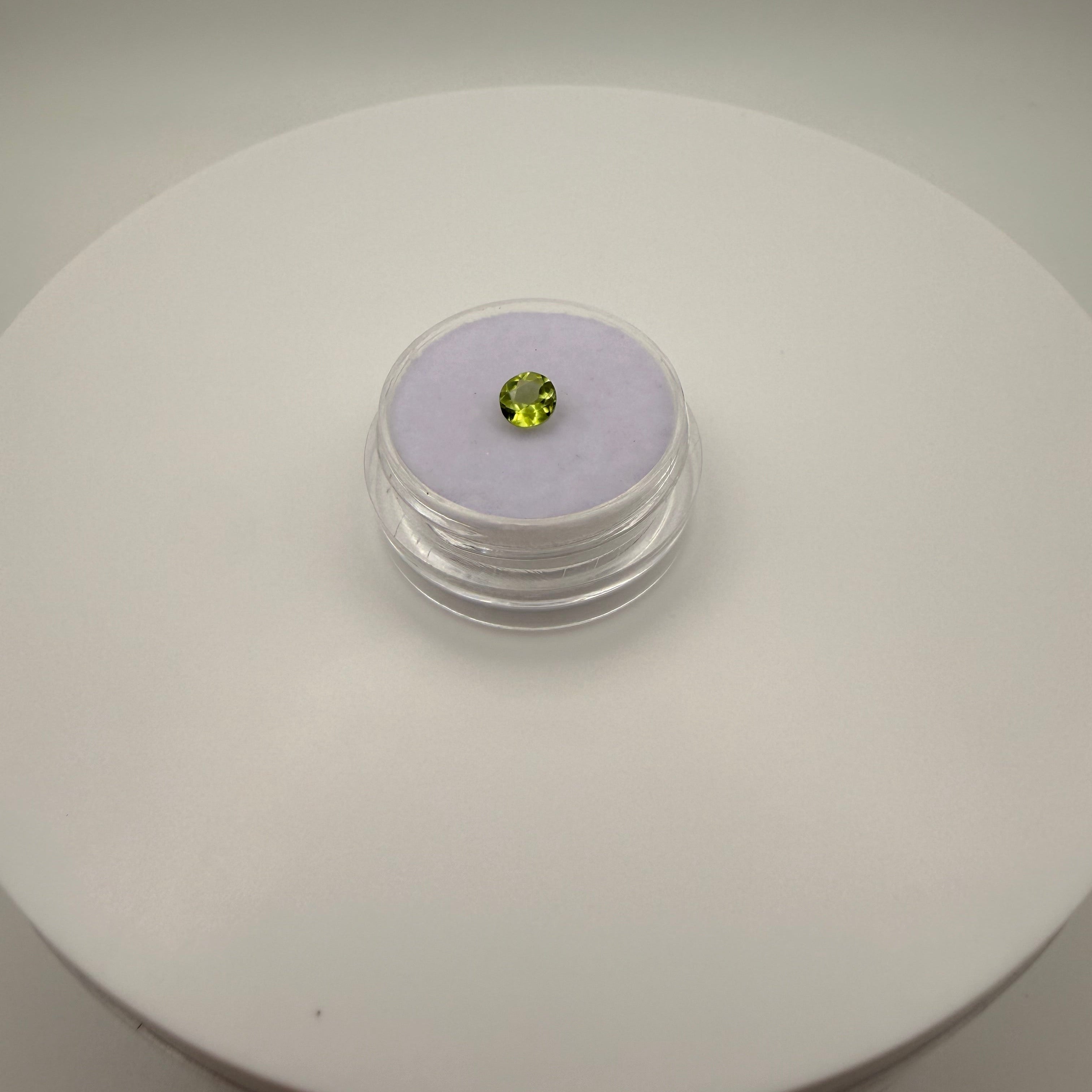 Macro view of a 0.45ct yellow-green peridot round faceted gemstone in a clear gem holder on a white circular surface.